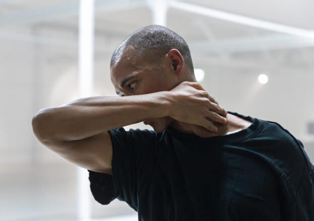 blackmilk at Stanley & Audrey Burton Theatre: A black man is pictured in motion with his profile central to the image, he is visible only from the waist up. The room is brightly lit and has white industrial features. He has short, dark, afro hair and is wearing an oversized black t-shirt. His right arm extends out in front of him, bending at the elbow, his hand reaches around, resting on the nape of his neck.