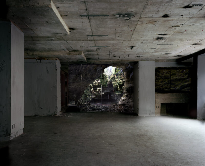 a grey, brutalist car park with a hole in the wall revealing a scene of nature- a grassy grotto and cave.