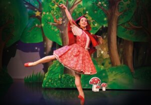 On a woodland background, a dancer wearing a red dress and a red cape balances on one foot.