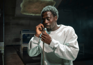 A man in a prison cell is stood holding a phone to his ear.  