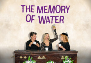 Text reads The Memory of Water. A image of three women in black sitting by a coffin, two with cups of tea, one in the middle raising a wineglass 