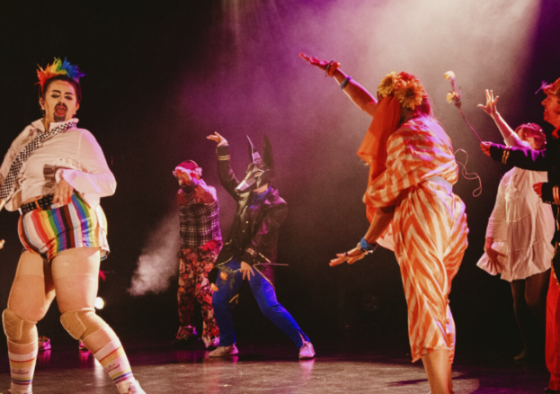 A number of performers dance on a stage wearing various costumes including rainbow hotpants and sunflower flower crowns