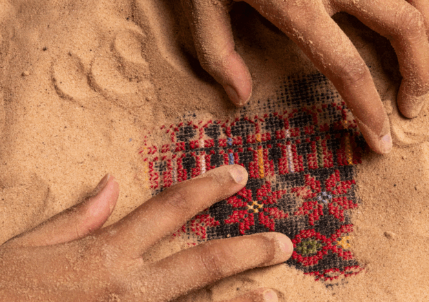 Two hands covered in sand are uncovering a piece of red fabric woven with flowers and a black and red repeating geometric pattern which is buried in the sand. 