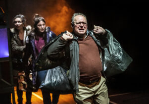 Three actors performing on stage, carrying black bin bags. 