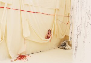 A doll with makeup peeks out of a hanging wall of butter yellow fabric. Red and black threads descend and cascade around the doll.