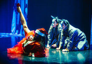 A festive drag queen lies sideways on a stage wearing a red santa hat, velvet santa jacket and red tutu. Raising one leg up towards the ceiling, wearing a glittery red high heel. Behind is two females dressed as donkeys.