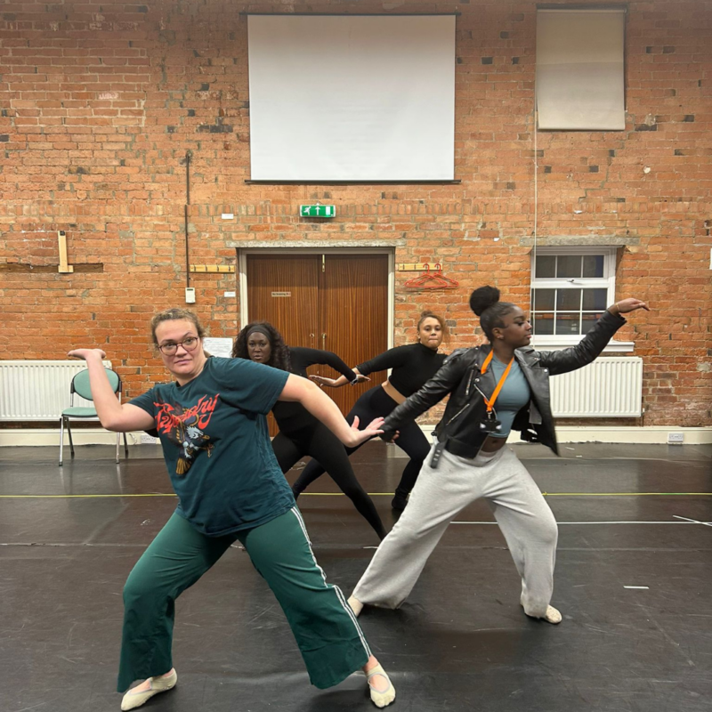 Four women rehearse a contemporary dance in a studio with brick walls, holding a sculptural pose with bent knees and outstretched arms, arranged in a layered formation