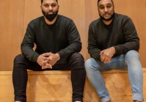 2 men of colour sat side by side in an atrium. Both wearing black tops, trainers and jeans.