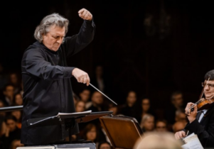 A white man with grey hair wearing all black, conducting an orchestra