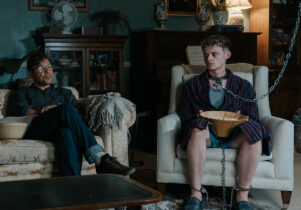 Scene from a film, the living room, darkly lit. A man watching TV with another man chained up.