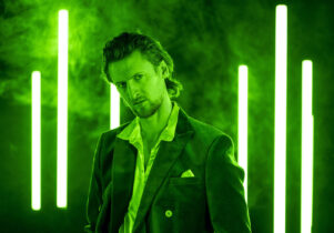 A man (Macbeth) stands in a dark studio lit in vivid green light, with vertical neon-green light bars glowing behind him through light fog. He wears a dark suit jacket over an open-collared shirt, one hand in his pocket, and looks directly at the camera with an intense, brooding expression.