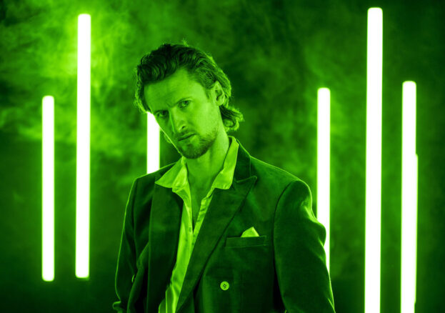 A man (Macbeth) stands in a dark studio lit in vivid green light, with vertical neon-green light bars glowing behind him through light fog. He wears a dark suit jacket over an open-collared shirt, one hand in his pocket, and looks directly at the camera with an intense, brooding expression.