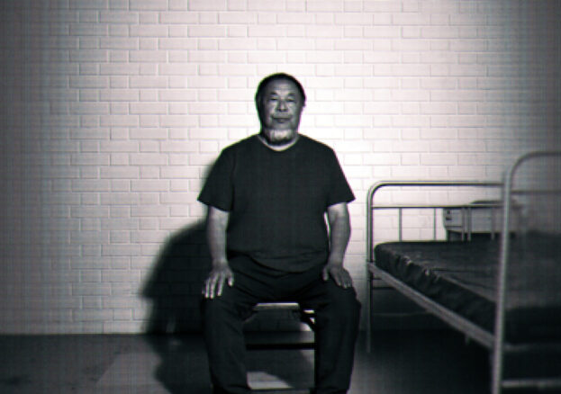 A black-and-white image of Ai Weiwei sitting upright on a small stool in a sparse, cell-like room. He faces the camera with his hands resting on his knees. Behind him is a white brick wall, and to his right there is a simple metal bed frame with a thin mattress. The lighting is stark, casting strong shadows on the wall and floor, giving the scene a stark, surveillance-like atmosphere