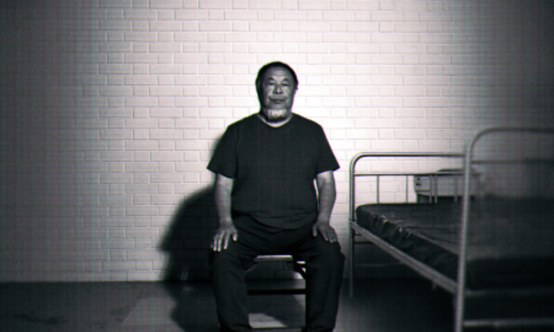 A black-and-white image of Ai Weiwei sitting upright on a small stool in a sparse, cell-like room. He faces the camera with his hands resting on his knees. Behind him is a white brick wall, and to his right there is a simple metal bed frame with a thin mattress. The lighting is stark, casting strong shadows on the wall and floor, giving the scene a stark, surveillance-like atmosphere