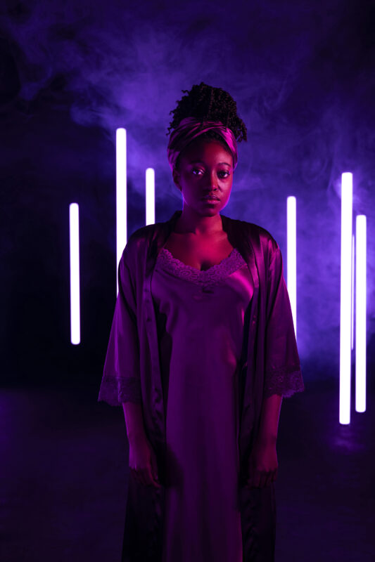 A woman (Lady Macbeth) stands facing forward in a dark, smoky space lit in purple and magenta tones, with vertical violet light bars glowing behind her. She wears a satin slip dress with lace trim and a loose robe, her hair wrapped in a headscarf, and gazes calmly at the camera with a composed, serious expression.