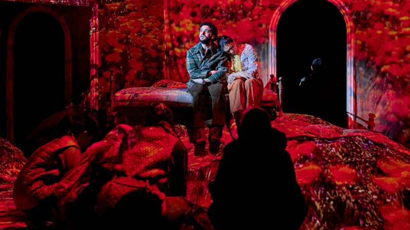 Two actors performing on stage. They are both sat on bed and there are red projections onto the stage.