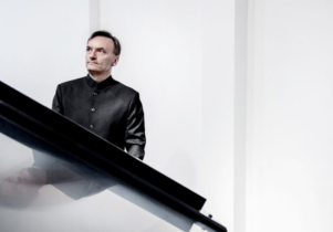 A white man with short dark hair wearing a black suit and standing behind a piano.