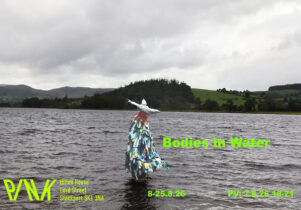 Poster for Bodies in Water. A figure wearing a multicoloured, fringed costume and silver star-shaped headpiece stands knee-deep in a lake beneath dark clouds, with green hills in the distance. Bright green text reads: “Bodies in Water”, “8–25.8.26” and “PV: 7.8.26, 18–21”. Bottom left shows the PINK logo and address: Hilton House, Lord Street, Stockport, SK1 3NA.
