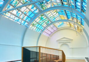 A large white room with a colourful lit ceiling above a wooden coffin shaped oversized sculpture.