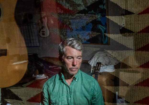 Bill Callahan in green shirt, looking down in a cluttered room with guitars