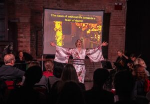 A singer in a white kimono with pink flowers sings with her arms spread wide, lit in warm red against a brick wall. Musicians play violin and cello beside her, the audience sits in the foreground, and a screen behind her reads: The dawn of artificial life demands a feast of death!