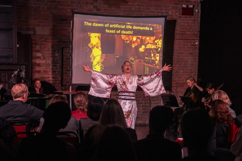 A singer in a white kimono with pink flowers sings with her arms spread wide, lit in warm red against a brick wall. Musicians play violin and cello beside her, the audience sits in the foreground, and a screen behind her reads: The dawn of artificial life demands a feast of death!