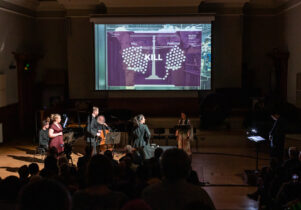 Singers and musicians on stage in a darkened concert hall, watched by a seated audience. A large screen above them shows a set of scales weighing KILL against FORGIVE, with the word KILL in large letters in the centre.